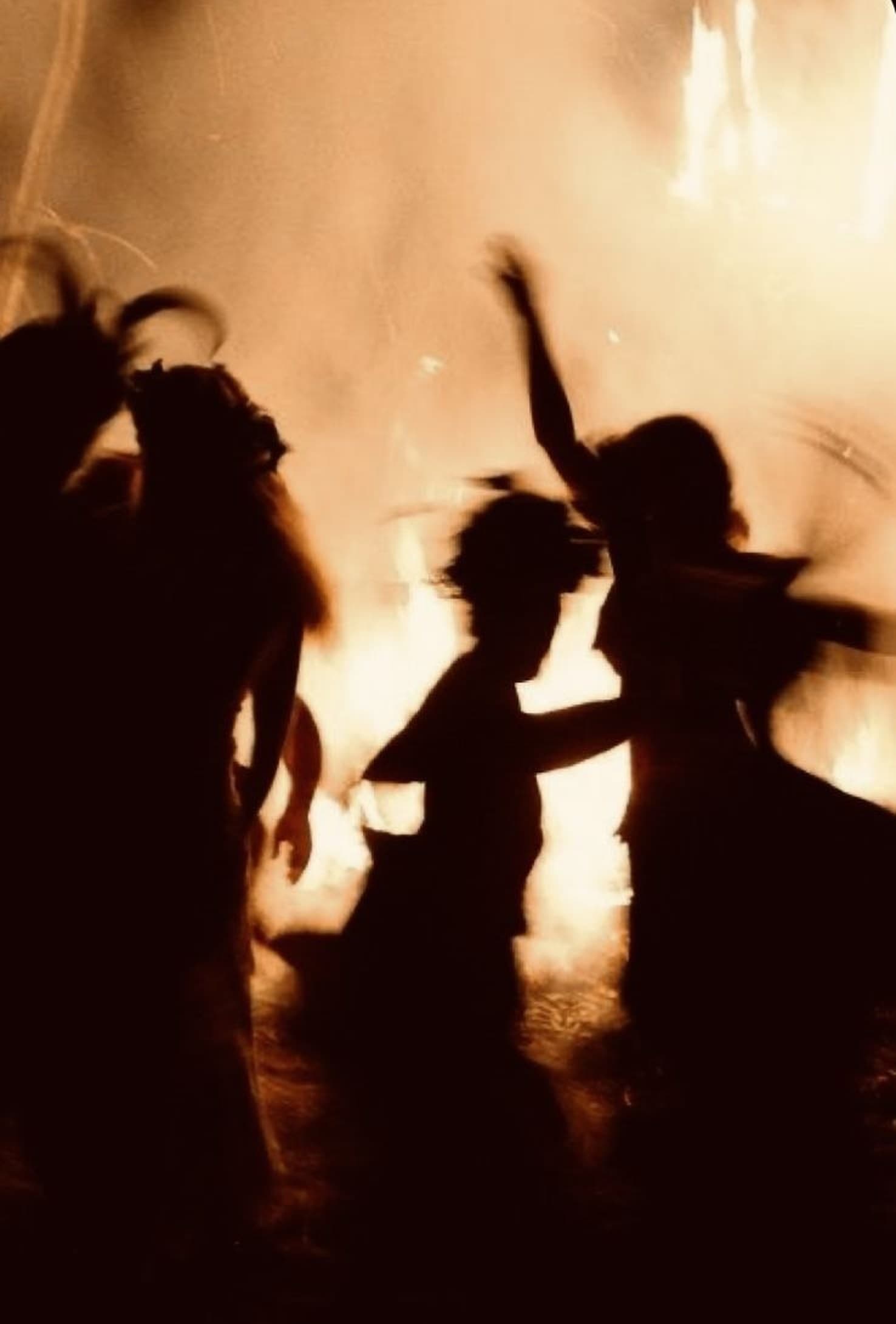 Silhouettes of women dancing around a bonfire at night, their movement blurred into trails against the firelight