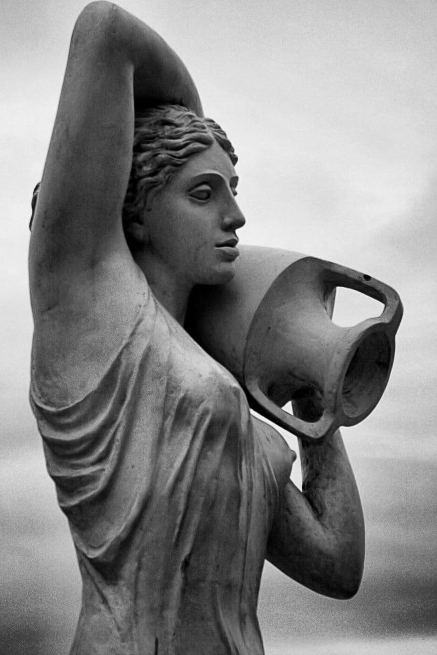 A stone statue of a woman raising an amphora to her shoulder, against an open sky